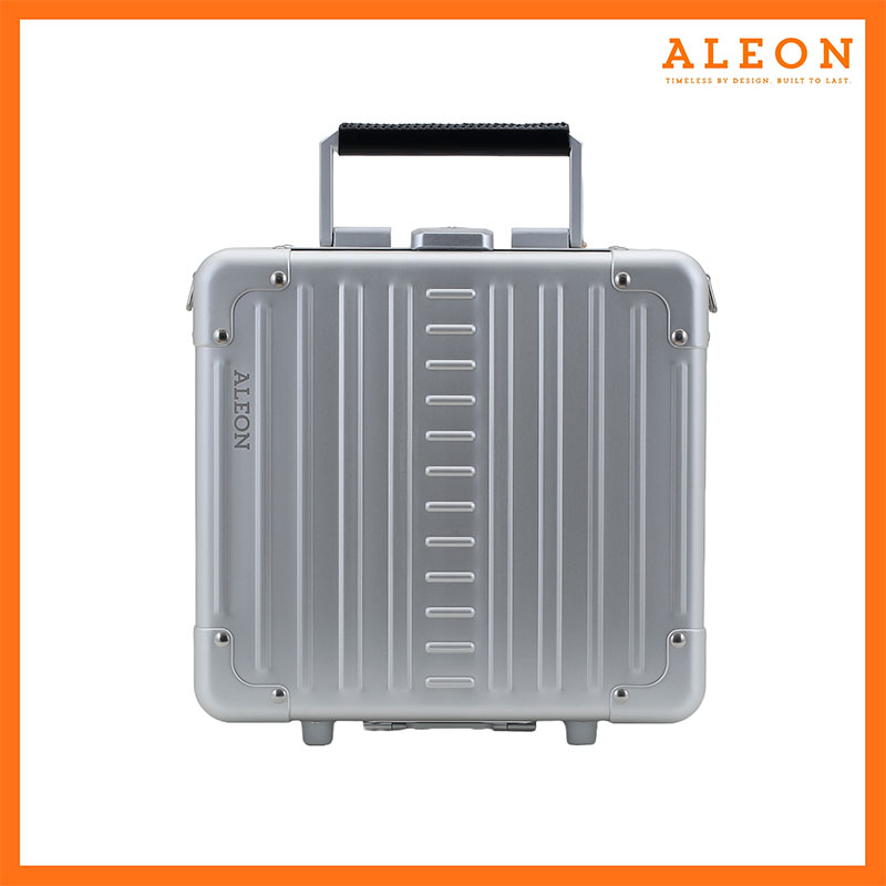 The image features a platinum silver ALEON Messenger Case with a sleek, modern design. Vertical ridges run across the aluminum exterior, reinforced with metal rivets at the corners for durability. A black handle sits at the top, complementing the case’s bold aesthetic.